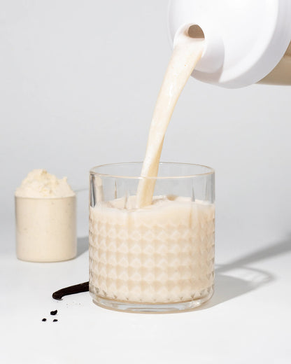 Glass filled with Grass-Fed Whey Protein drink being poured from a container, showcasing a creamy texture.